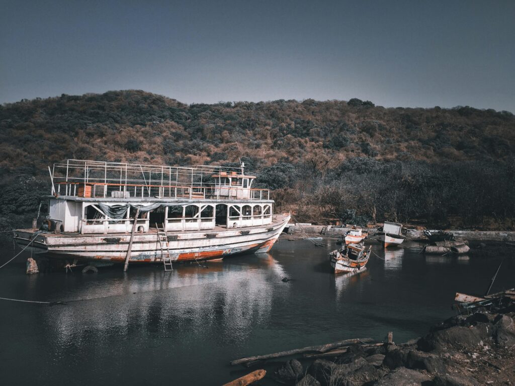 An old, rustic boat docked by lush coastal hills in Navi Mumbai, India.