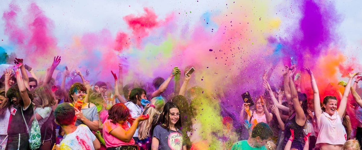 A joyful crowd covered in vivid colors celebrates a festival outdoors with flying powder.